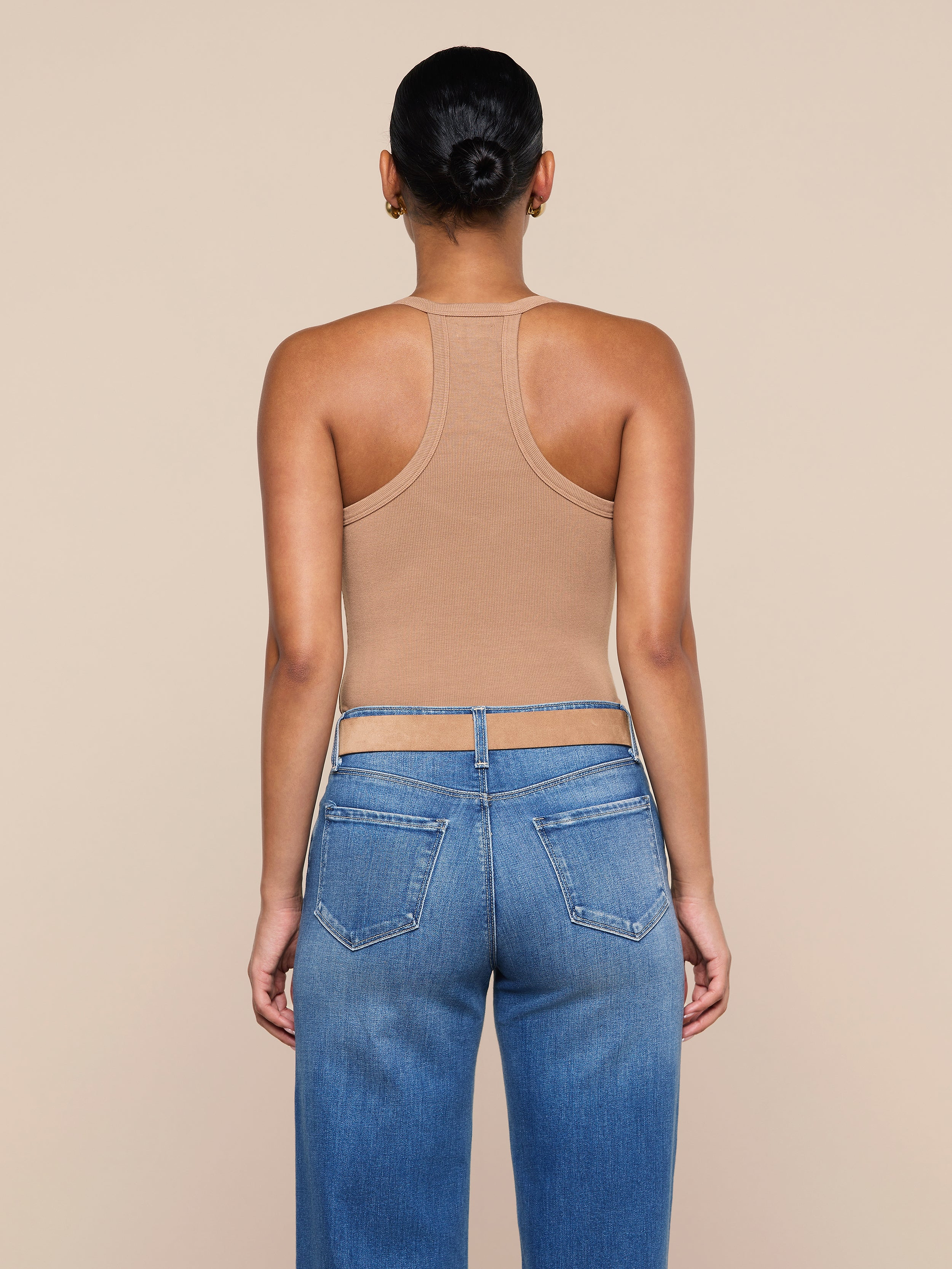 A person with their hair in a bun, wearing the Wrenna Racerback Bodysuit in tan rib fabric and blue jeans, stands facing away from the camera against a plain, light background.