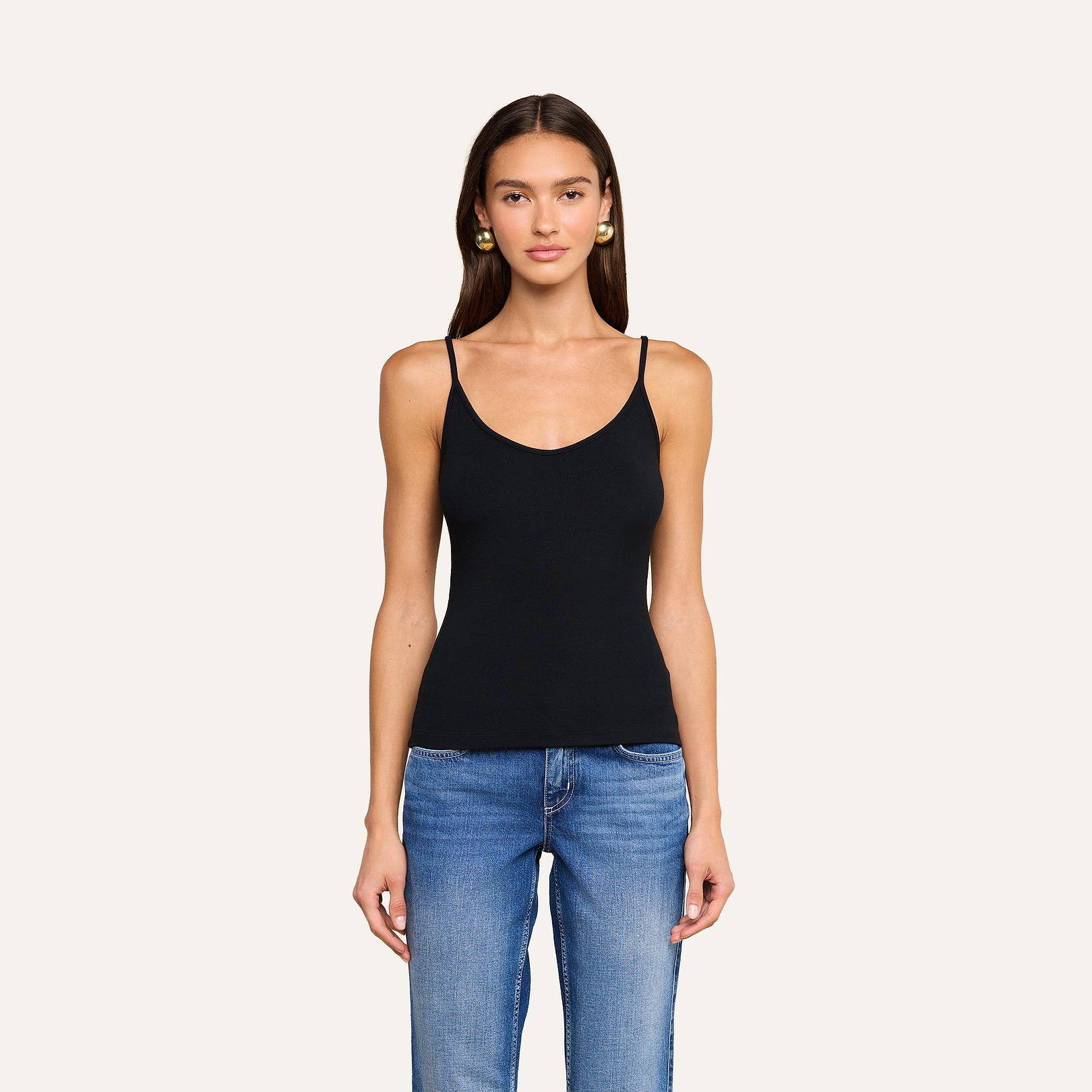 A woman with long brown hair, wearing a black ribbed Lunara Camisole and blue jeans, stands facing forward against a plain light background.