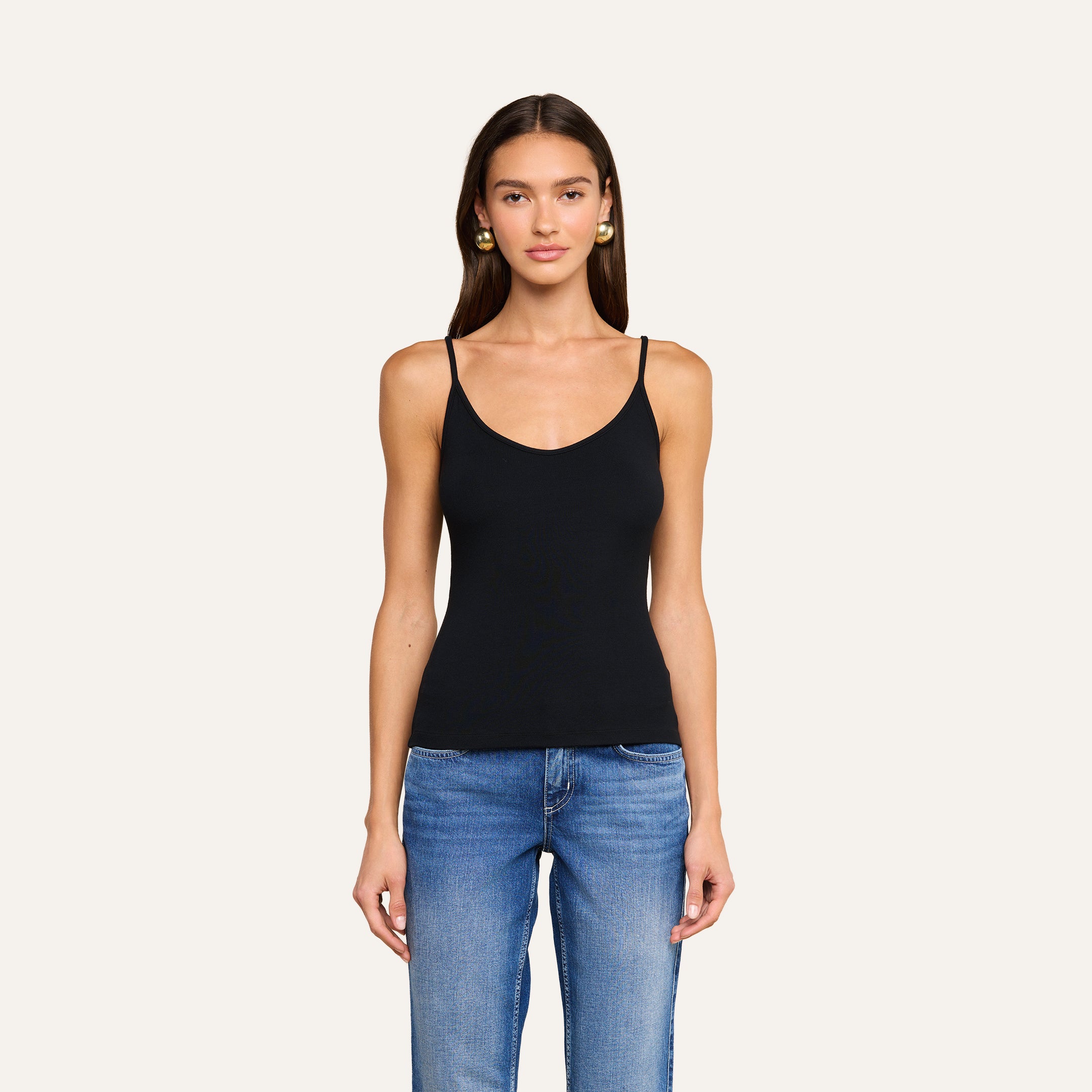 A woman with long brown hair, wearing a black ribbed Lunara Camisole and blue jeans, stands facing forward against a plain light background.