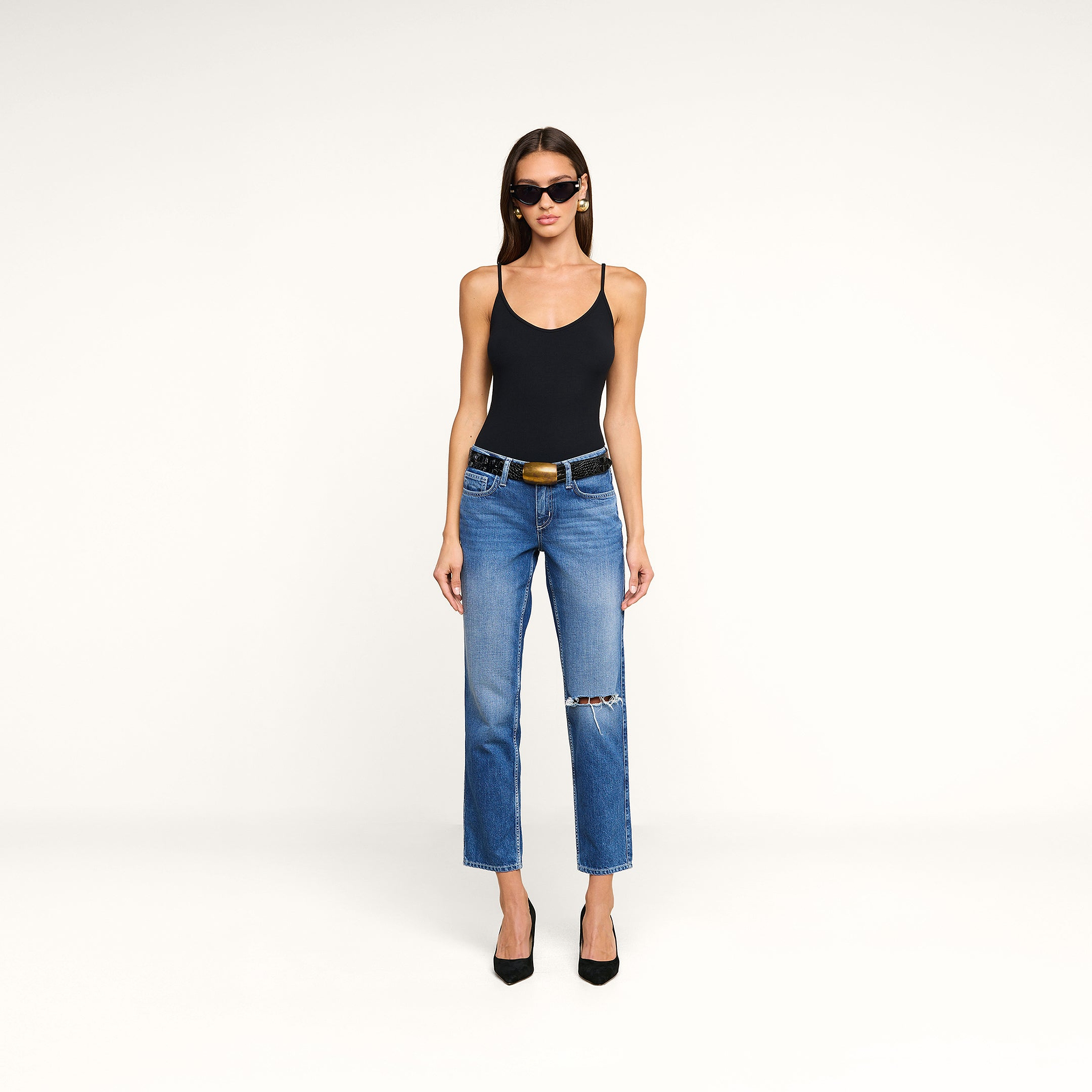 Woman wearing a black Lunara Camisole made from soft micro modal ribbed fabric, blue ripped jeans with a belt, black high heels, and sunglasses, standing against a plain white background.