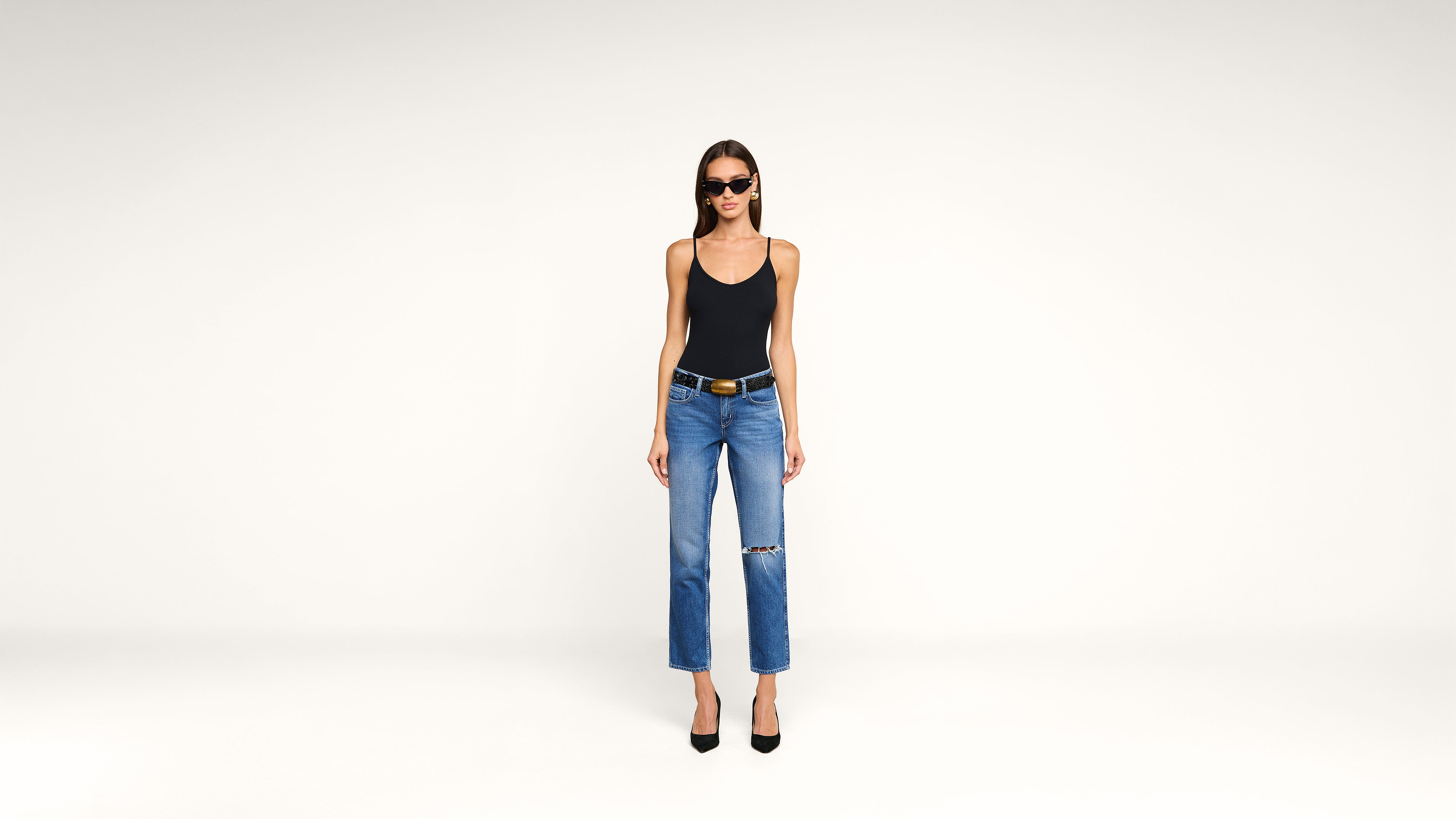 Woman wearing sunglasses, a black tank top, blue jeans with a knee rip, a black belt, and black heels stands against a plain white background.