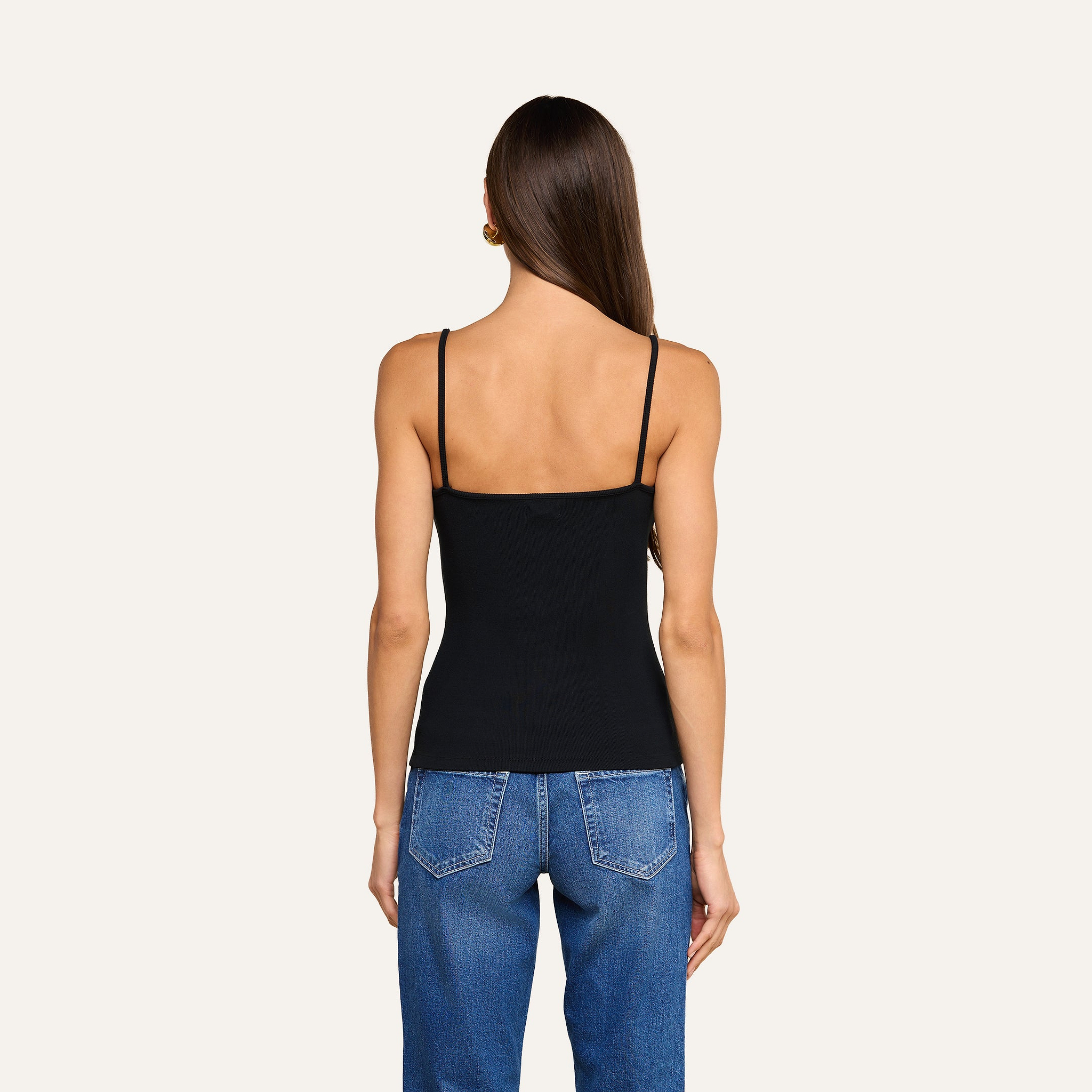 A woman stands with her back to the camera, wearing the Lunara Camisole in black ribbed fabric and blue jeans against a plain, light background.