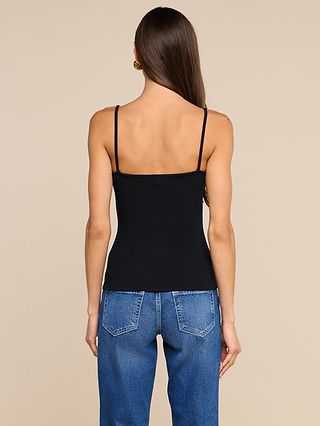 A woman with long brown hair, dressed in a black Lunara Camisole made from soft ribbed fabric and blue jeans, stands facing away from the camera against a beige background.