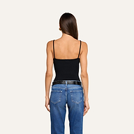 A woman stands facing away, wearing the Lunara Camisole—a black ribbed fabric spaghetti strap top—paired with blue jeans and a black belt. Her long brown hair is straight and loose, and her arms rest relaxed at her sides against a plain light background.