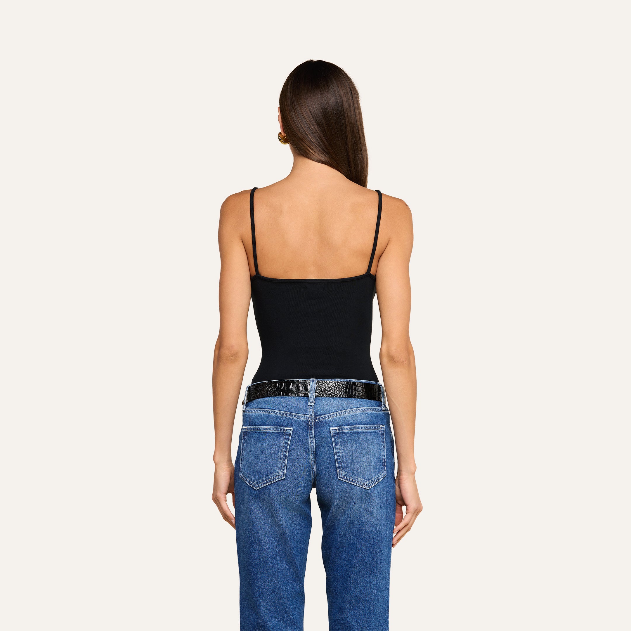 A woman stands facing away, wearing the Lunara Camisole—a black ribbed fabric spaghetti strap top—paired with blue jeans and a black belt. Her long brown hair is straight and loose, and her arms rest relaxed at her sides against a plain light background.