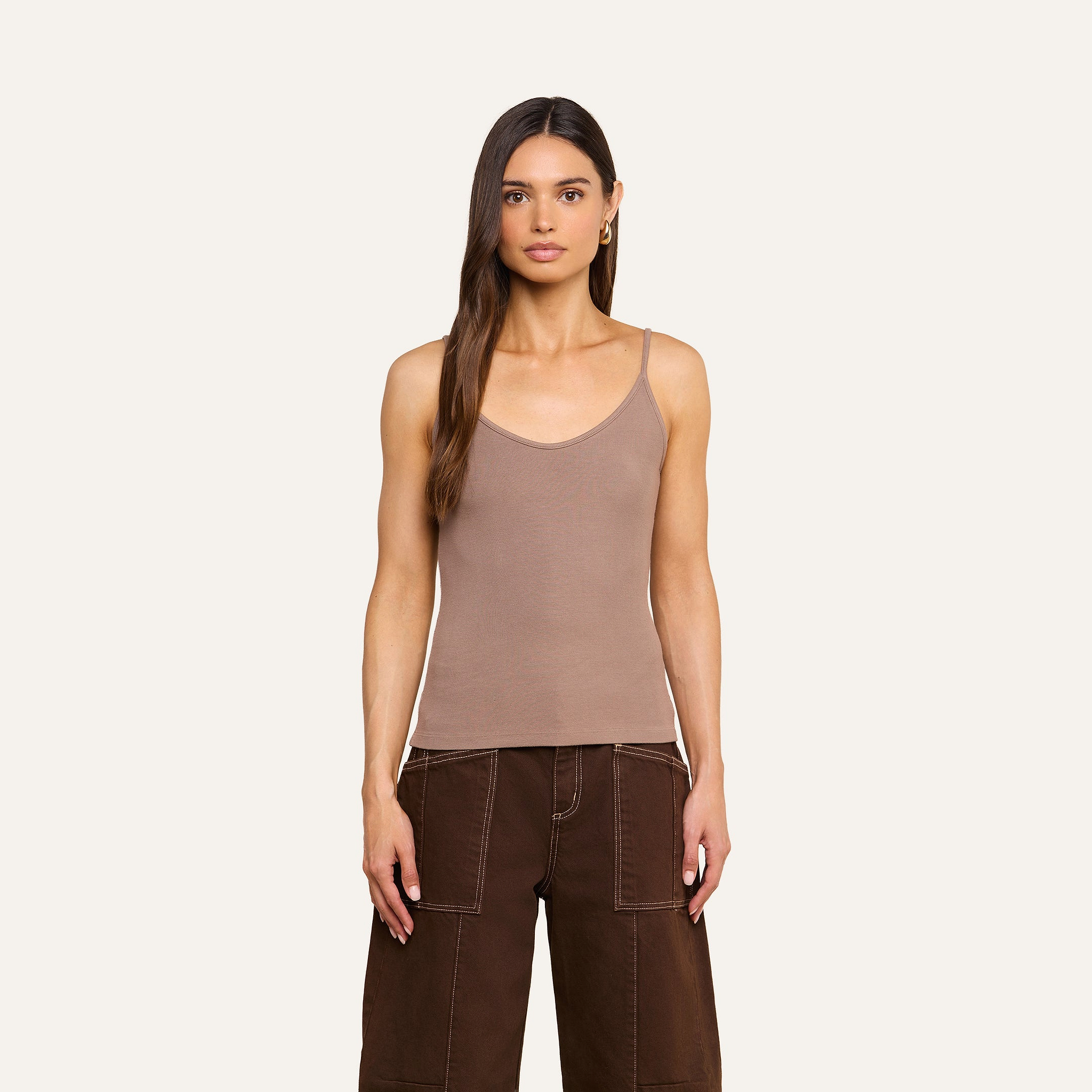 A woman with long brown hair stands facing forward against a plain light background, wearing a beige Lunara Camisole crafted from ribbed fabric and brown wide-leg pants.