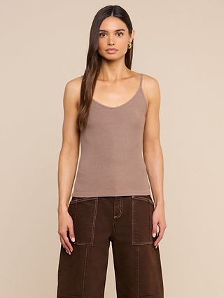 A woman with long brown hair wears a taupe Lunara Camisole crafted from ribbed fabric and brown wide-leg pants, standing in front of a plain beige background.