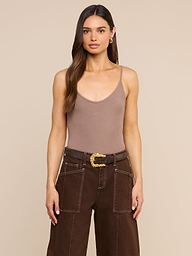 A woman with long brown hair wears a Lunara Camisole crafted from soft micro modal ribbed fabric, paired with dark brown pants featuring white stitching and a decorative belt, standing against a plain light background.
