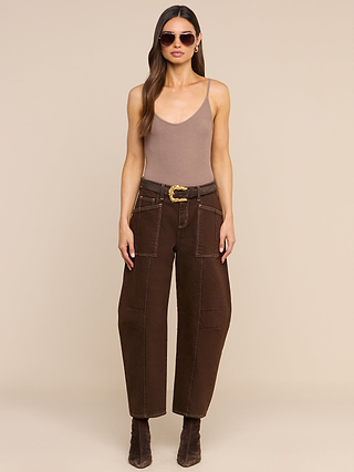Woman wearing sunglasses, a taupe Lunara Camisole crafted from micro modal ribbed fabric, brown belted pants with large pockets, and brown ankle boots stands against a beige background.
