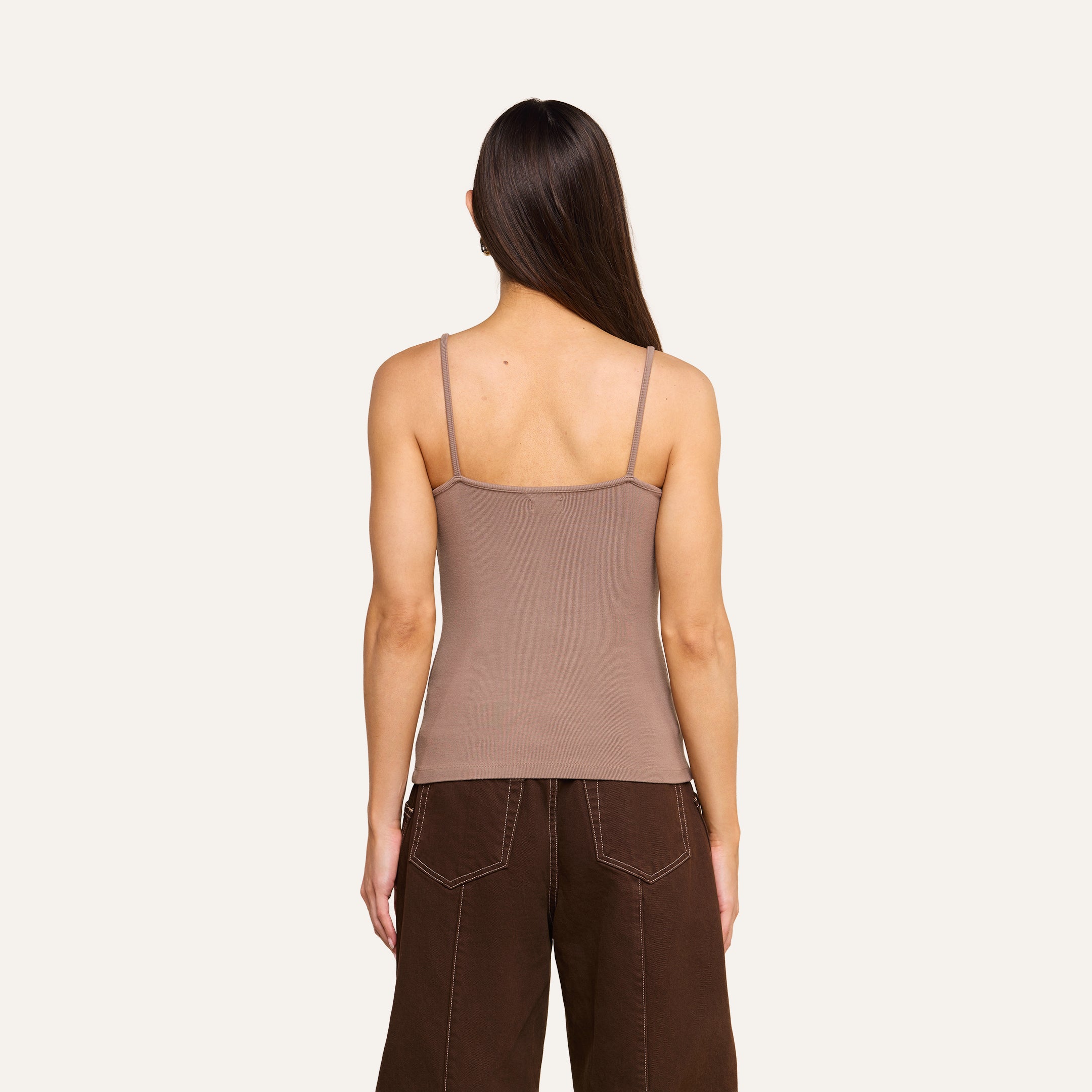 A woman with long brown hair is shown from the back, wearing a taupe Lunara Camisole made from ribbed fabric and dark brown pants against a plain light background.