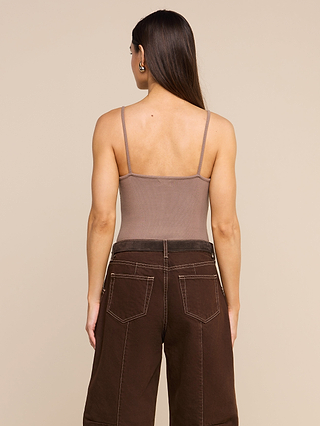 A woman with long brown hair, wearing a taupe micro modal Lunara Camisole and dark brown wide-leg jeans, stands facing away from the camera against a plain beige background.