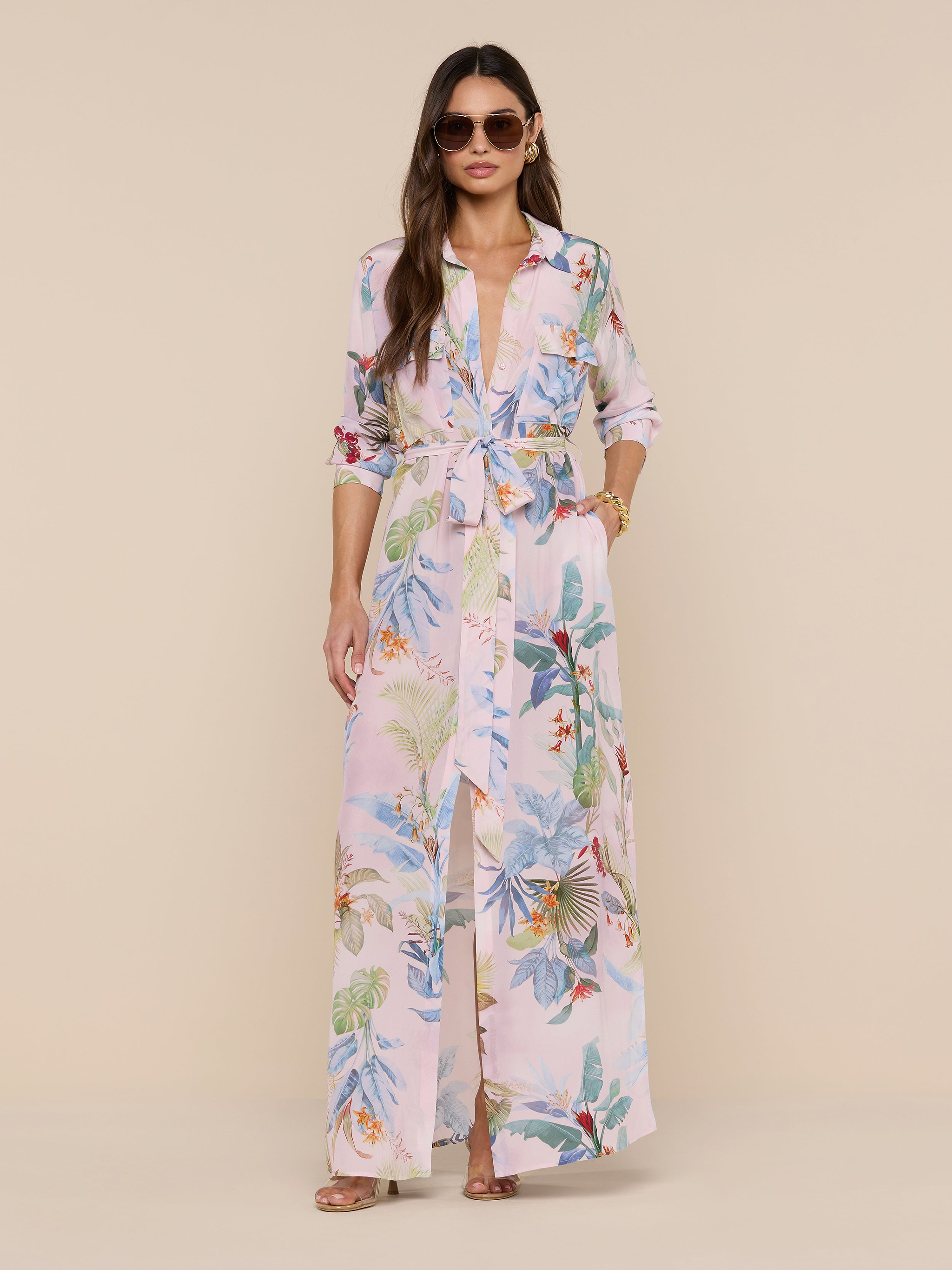 Woman in sunglasses and the Cameron Silk Shirt Dress—featuring a belted waist and rolled-up sleeves—stands against a beige background with loose hair, bracelets, and rings.