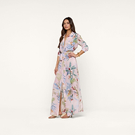 A woman wears the Cameron Silk Shirt Dress—a long, flowy silk crepe de chine with a light pink floral pattern and belted waist—paired with nude heeled sandals and gold bracelets, set against a plain beige background.