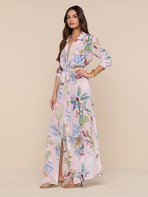 A woman wears the Cameron Silk Shirt Dress—a long, flowy silk crepe de chine with a light pink floral pattern and belted waist—paired with nude heeled sandals and gold bracelets, set against a plain beige background.