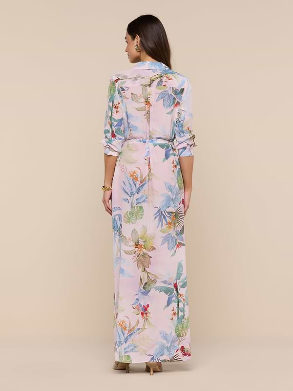 A woman stands facing away, wearing the Cameron Silk Shirt Dress—a long, light pink floral crepe de chine dress with long sleeves and a belted waist—paired with beige heels against a plain beige background.