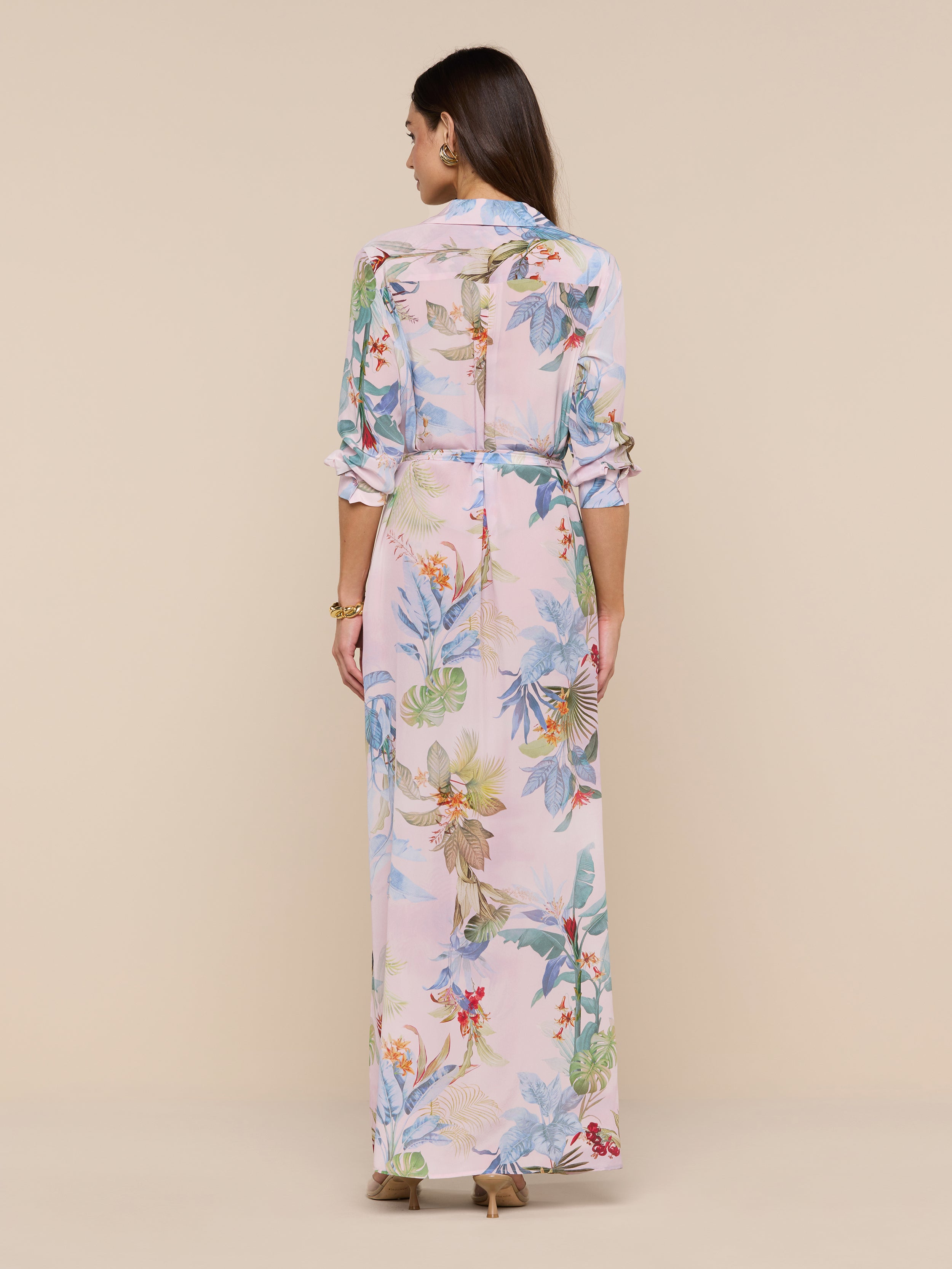A woman stands facing away, wearing the Cameron Silk Shirt Dress—a long, light pink floral crepe de chine dress with long sleeves and a belted waist—paired with beige heels against a plain beige background.