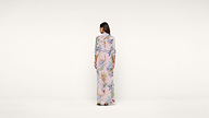 A woman stands facing away, wearing a long, light-colored dress with a colorful floral pattern, against a plain white background.