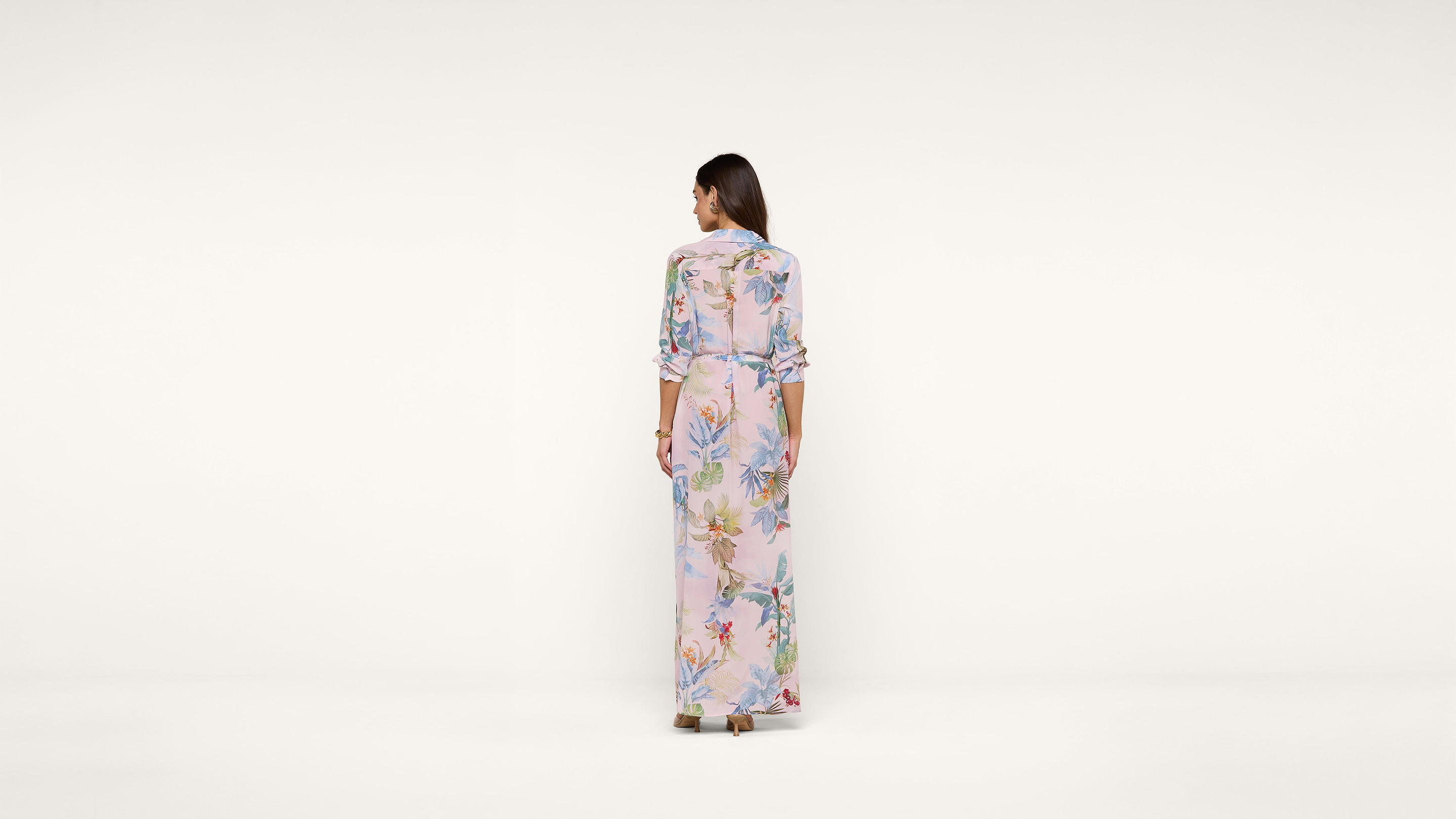 A woman stands facing away, wearing a long, light-colored dress with a colorful floral pattern, against a plain white background.