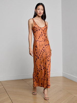 A woman with long, straight hair wears the L'AGENCE Akiya Satin Maxi Dress—a burnt orange, bias-cut dress with a geometric pattern. She pairs it with heeled sandals and poses in a minimal room, one hand relaxed by her side.