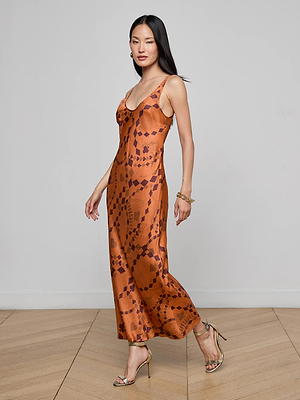 A woman in the L'AGENCE Akiya Satin Maxi Dress stands on a wooden floor against a light gray wall. The bias-cut burnt orange dress features a geometric pattern. She pairs it with gold heels, bracelets, earrings, and wears her long hair straight.