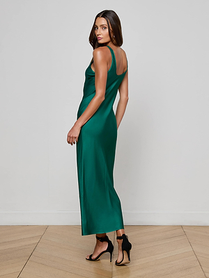A woman wears the Akiya Satin Maxi Dress, a sleeveless emerald green gown, paired with black high-heeled sandals on a wooden floor, glancing over her shoulder against a simple light background.