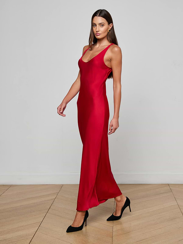 A woman with long brown hair wears the L'AGENCE Akiya Satin Maxi Dress, a sleeveless red, floor-length bias-cut design, paired with black heels as she walks on a light wood floor against a white wall, gazing ahead neutrally.