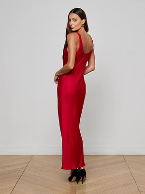 A woman with long brown hair models the L'AGENCE Akiya Satin Maxi Dress in red, paired with black heels, standing on a wooden floor and posing elegantly with her back turned against a white wall.