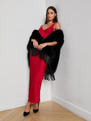 A woman in the L'AGENCE Akiya Satin Maxi Dress and black heels leans against a white wall, draped in a black fringe fur shawl, standing on a wooden floor.
