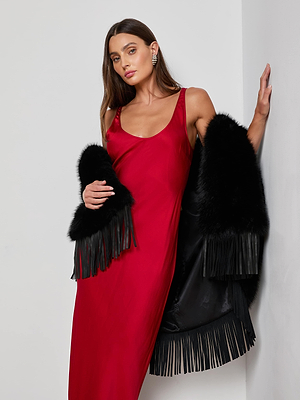 A woman with long brown hair wears the L'AGENCE Akiya Satin Maxi Dress, styled with a black fur-trimmed fringe jacket, standing against a white wall and exuding confidence—an all-season essential.
