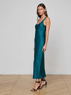 A woman poses sideways on a wooden floor in front of a white wall, wearing the L'AGENCE Akiya Satin Maxi Dress in teal with thin straps and black high-heeled sandals, looking toward the camera.