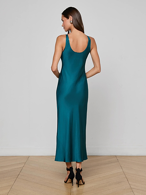 A woman with long brown hair stands on a light wood floor, wearing the L'AGENCE Akiya Satin Maxi Dress in teal and black high-heeled sandals, facing away from the camera against a plain white wall.