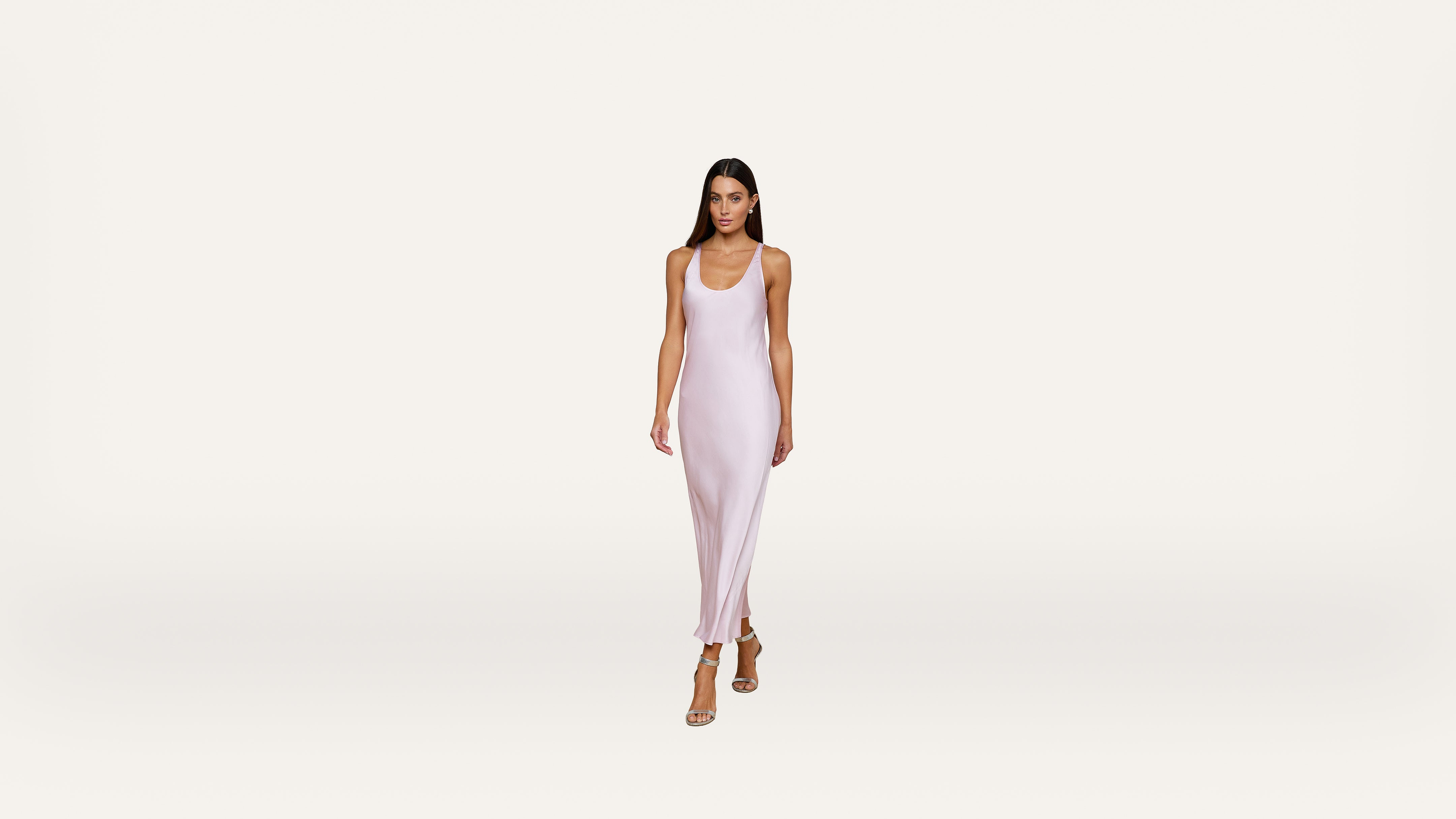A woman wearing a light pink, sleeveless, ankle-length dress and beige heels walks forward against a plain white background.