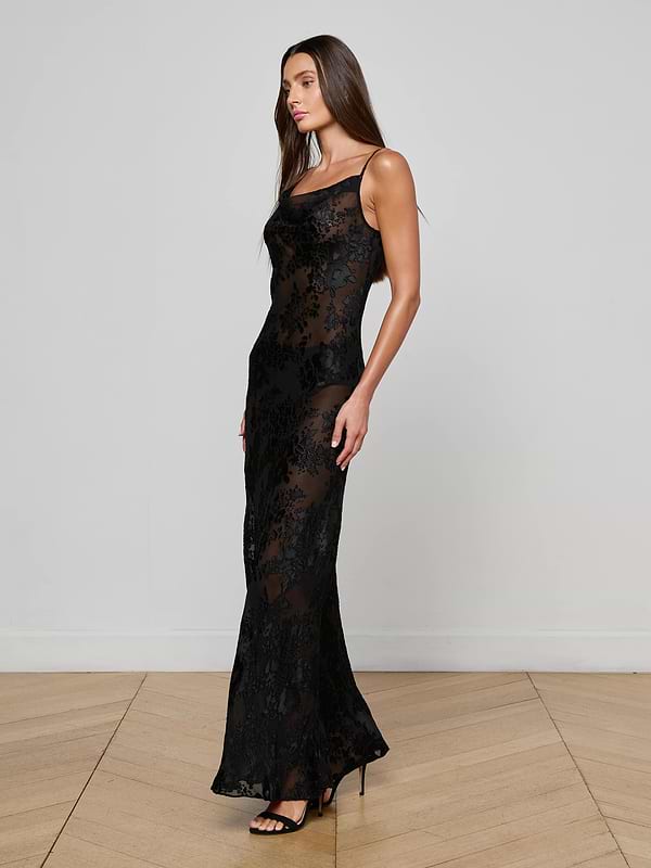 A woman stands sideways on a wooden floor against a white wall, wearing the L'AGENCE Christine Burnout Satin Dress—a long, fitted, sleeveless black dress with sheer floral lace overlay—and rests one arm by her side.