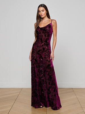 A woman with long brown hair poses on a wooden floor against a light gray wall, wearing the L'AGENCE Christine Burnout Velvet Dress—a sleeveless, floor-length dress with a deep purple floral pattern.