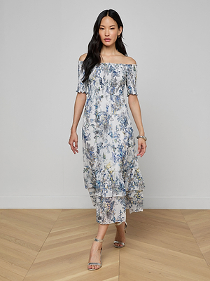 A woman in the Oriel Off-Shoulder Dress, featuring a blue and white floral print and ruffle hem, walks indoors on a wooden floor with her hair down and silver sandals.