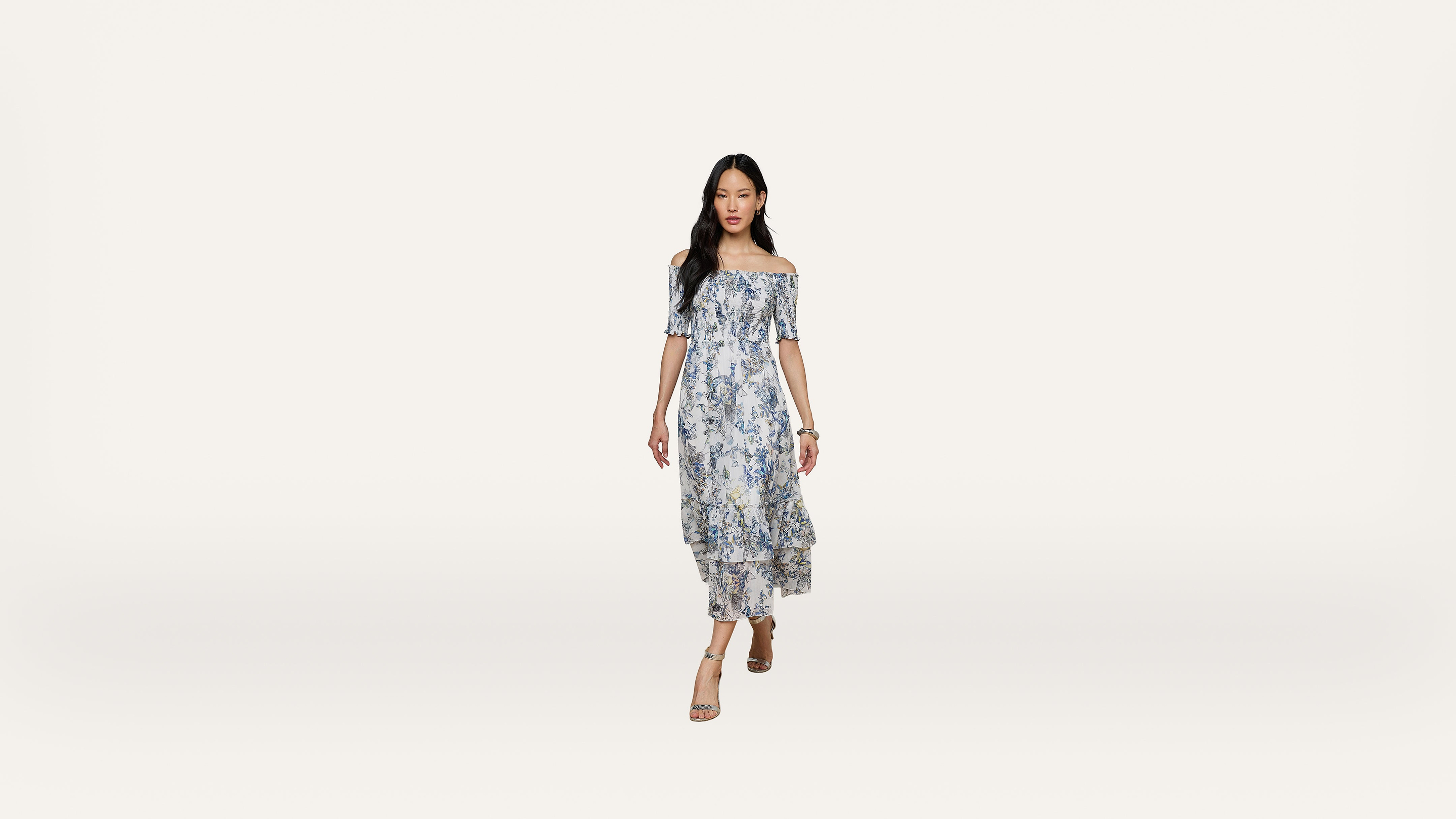 A woman in an off-shoulder, blue floral dress and sandals walks forward against a plain white background.