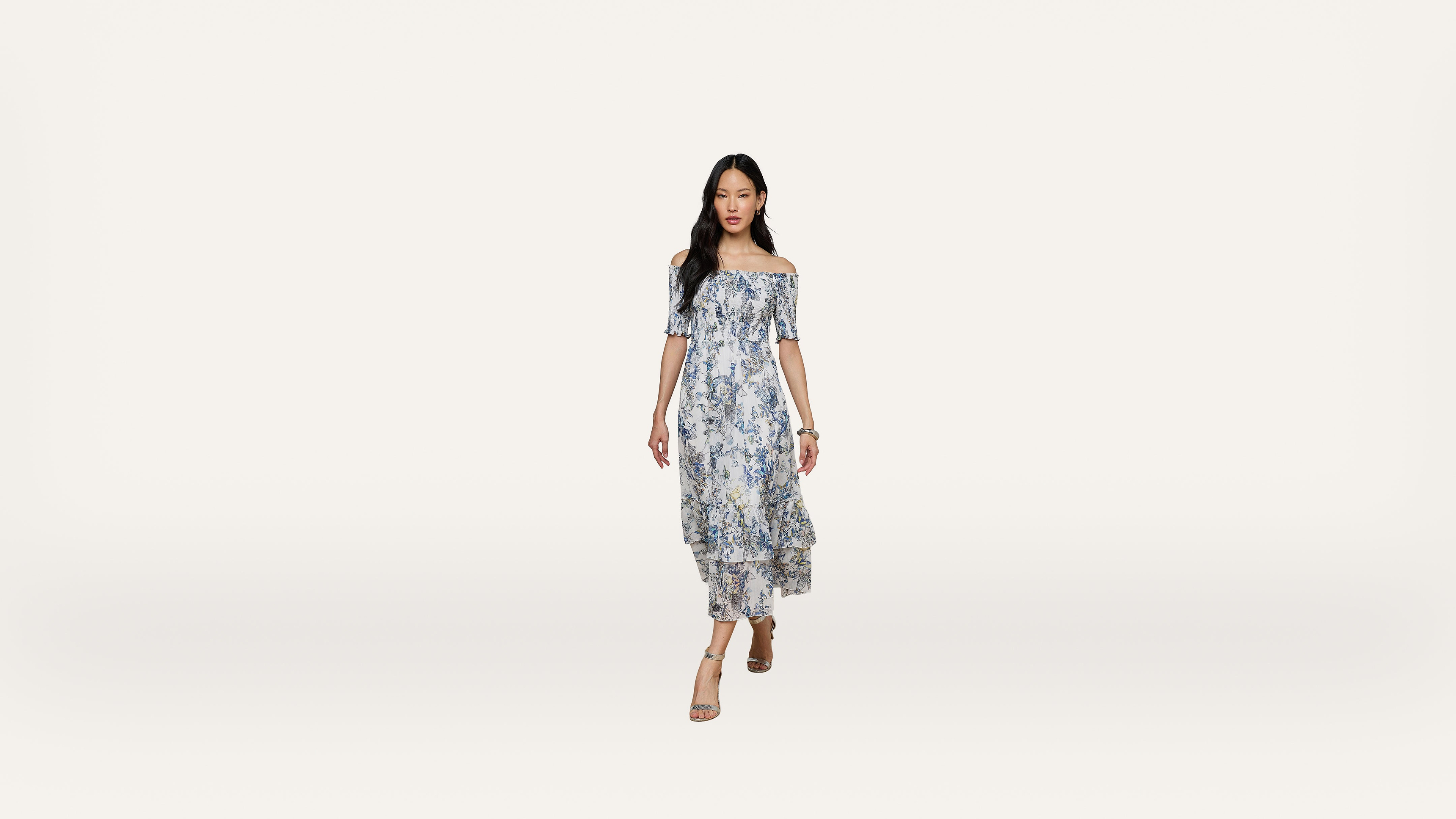A woman in an off-shoulder, blue floral dress and sandals walks forward against a plain white background.
