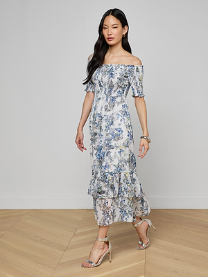 A woman with long dark hair walks on a light wood floor wearing the Oriel Off-Shoulder Dress, featuring ankle-length ruffles, short sleeves, and a butterfly print, paired with silver high-heeled sandals and a bracelet.