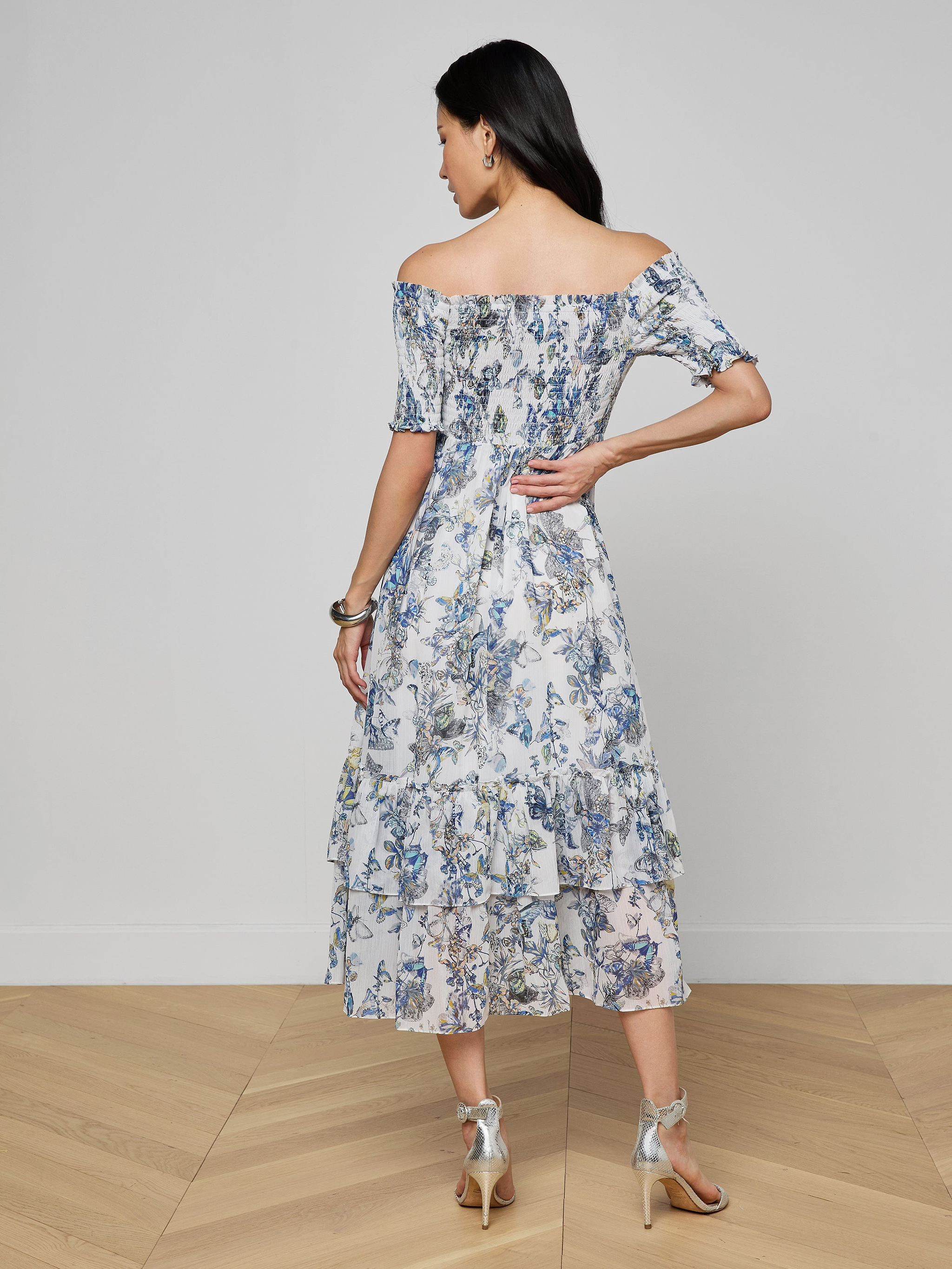 A woman with long dark hair stands on a wooden floor, facing away, wearing the Oriel Off-Shoulder Dress featuring floral and butterfly prints. She pairs it with silver high-heeled sandals, hoop earrings, and a bangle.