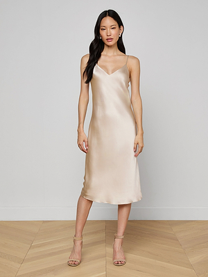 A woman stands indoors against a light wall and wooden floor, wearing the Seridie Silk Slip Dress in champagne with thin straps and nude heeled sandals.