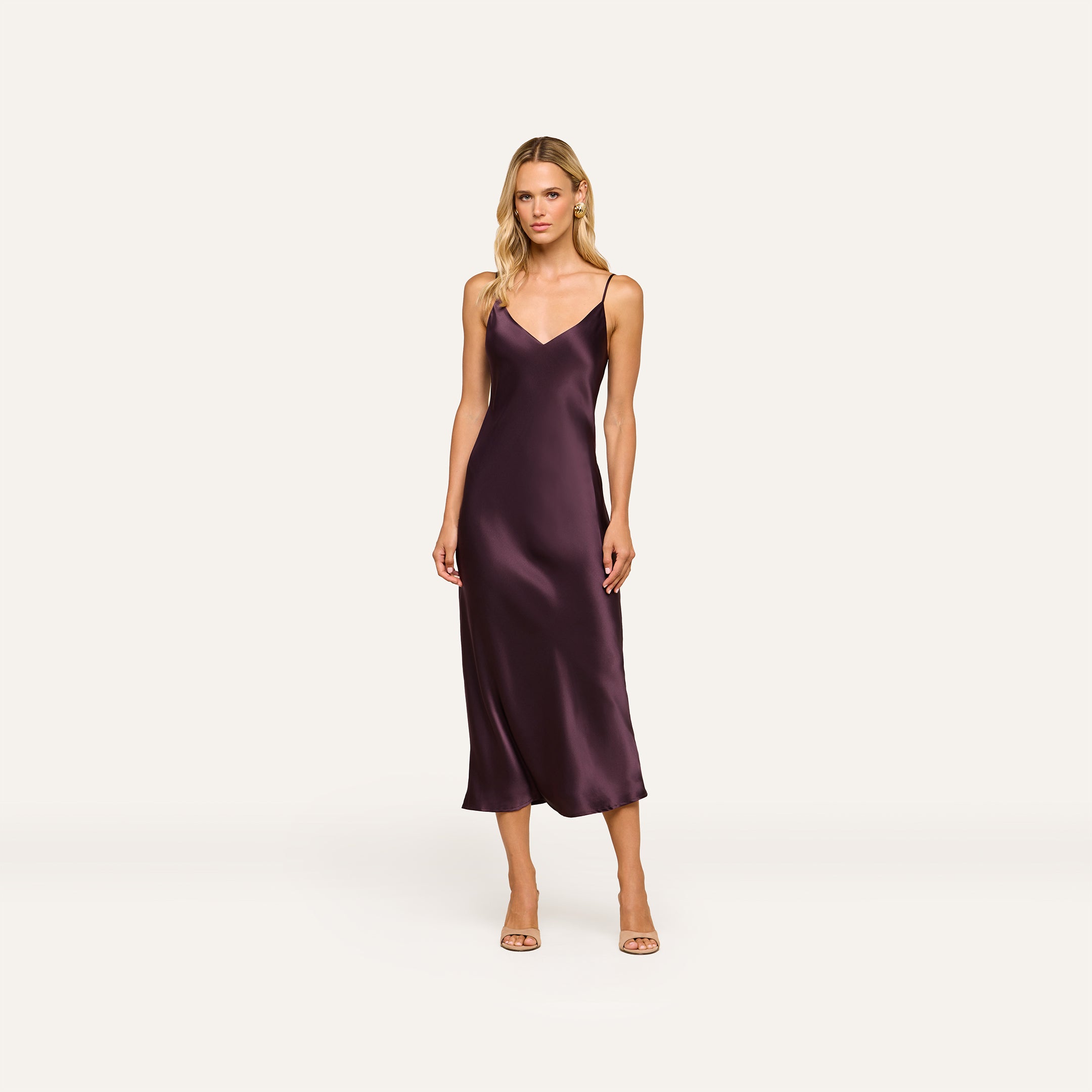 Wearing the Seridie Silk Slip Dress in deep purple and nude heels, a woman stands against a plain, light background, looking toward the camera with a neutral expression.