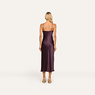 A woman with long blonde hair stands facing away, wearing the Seridie Silk Slip Dress—a sleeveless, dark purple bias-cut evening dress—paired with beige heels against a plain light background.