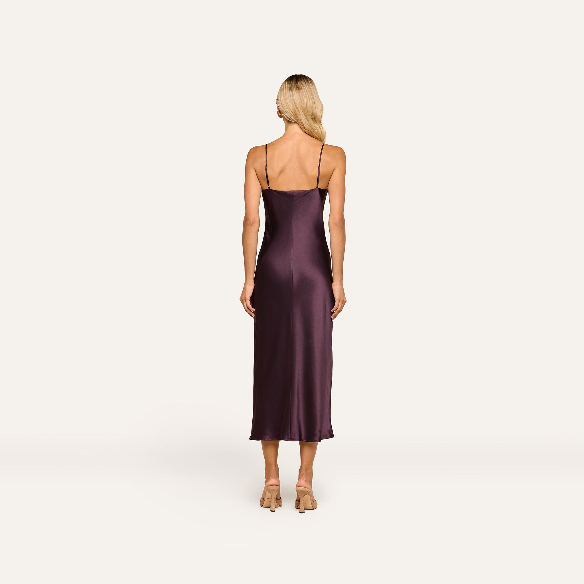 A woman with long blonde hair stands facing away, wearing the Seridie Silk Slip Dress—a sleeveless, dark purple bias-cut evening dress—paired with beige heels against a plain light background.