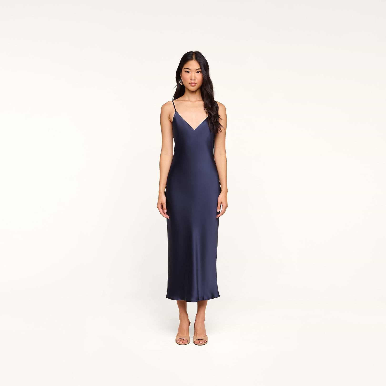 A woman wearing the Seridie Silk Slip Dress—a navy blue, sleeveless, V-neck midi dress with thin straps—stands against a plain white background, facing forward with a neutral expression, making it an elegant choice for a wedding guest.