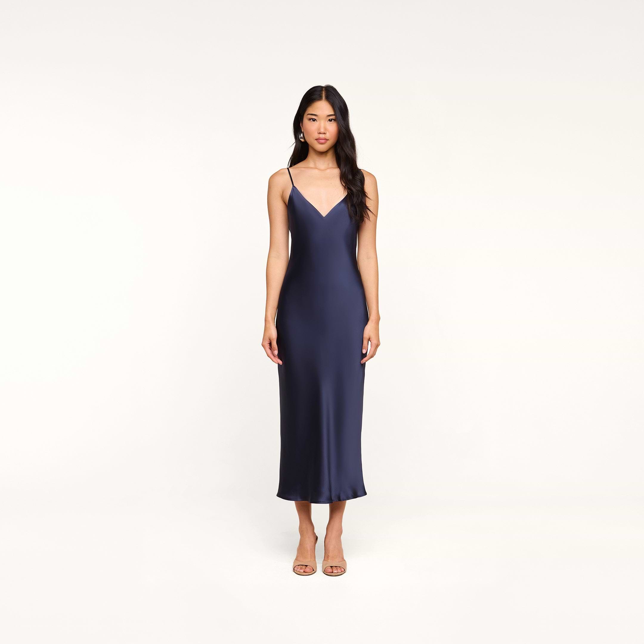 A woman wearing the Seridie Silk Slip Dress—a navy blue, sleeveless, V-neck midi dress with thin straps—stands against a plain white background, facing forward with a neutral expression, making it an elegant choice for a wedding guest.