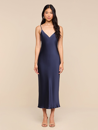 A woman stands facing forward against a plain beige background, wearing the Seridie Silk Slip Dress—a navy blue midi dress featuring a sleeveless, V-neck satin slip silhouette with delicate spaghetti straps—paired with nude heels. This elegant ensemble is perfect as a wedding guest dress.
