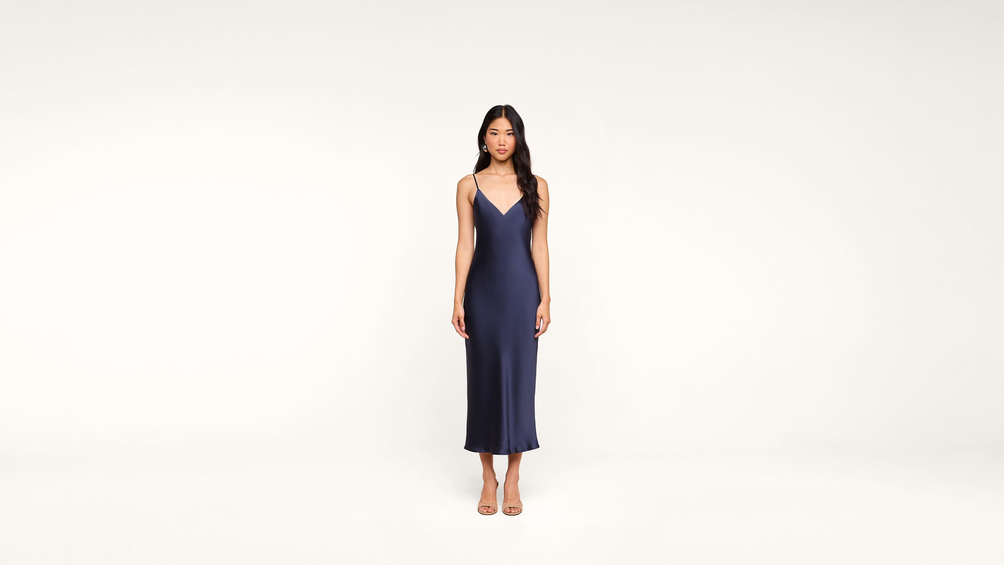 A woman stands against a plain white background, wearing a sleeveless, navy blue, midi-length dress with spaghetti straps and open-toe heels.