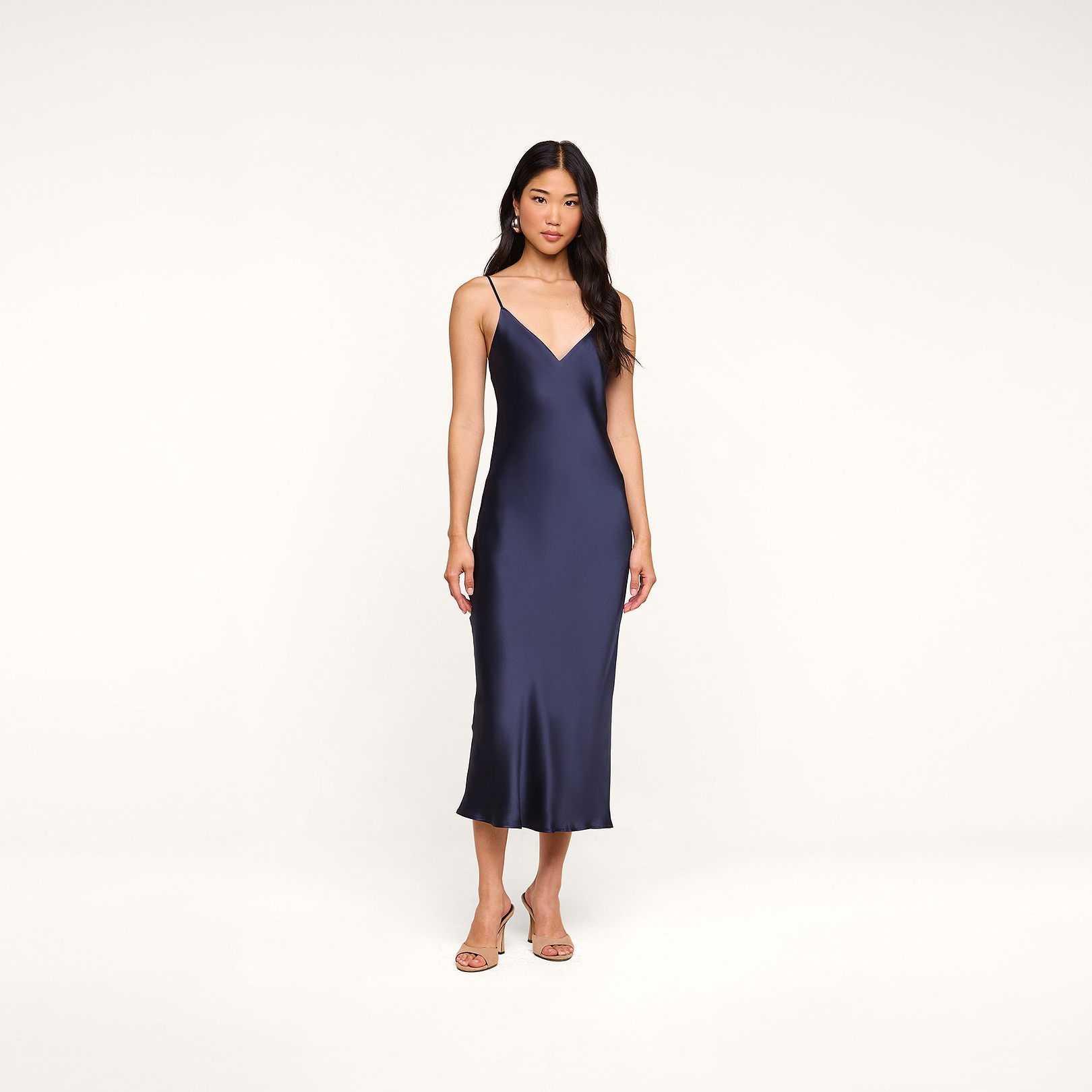 A woman stands against a plain white background wearing the Seridie Silk Slip Dress in navy blue with thin straps, paired with beige heeled sandals—an elegant choice for a wedding guest.