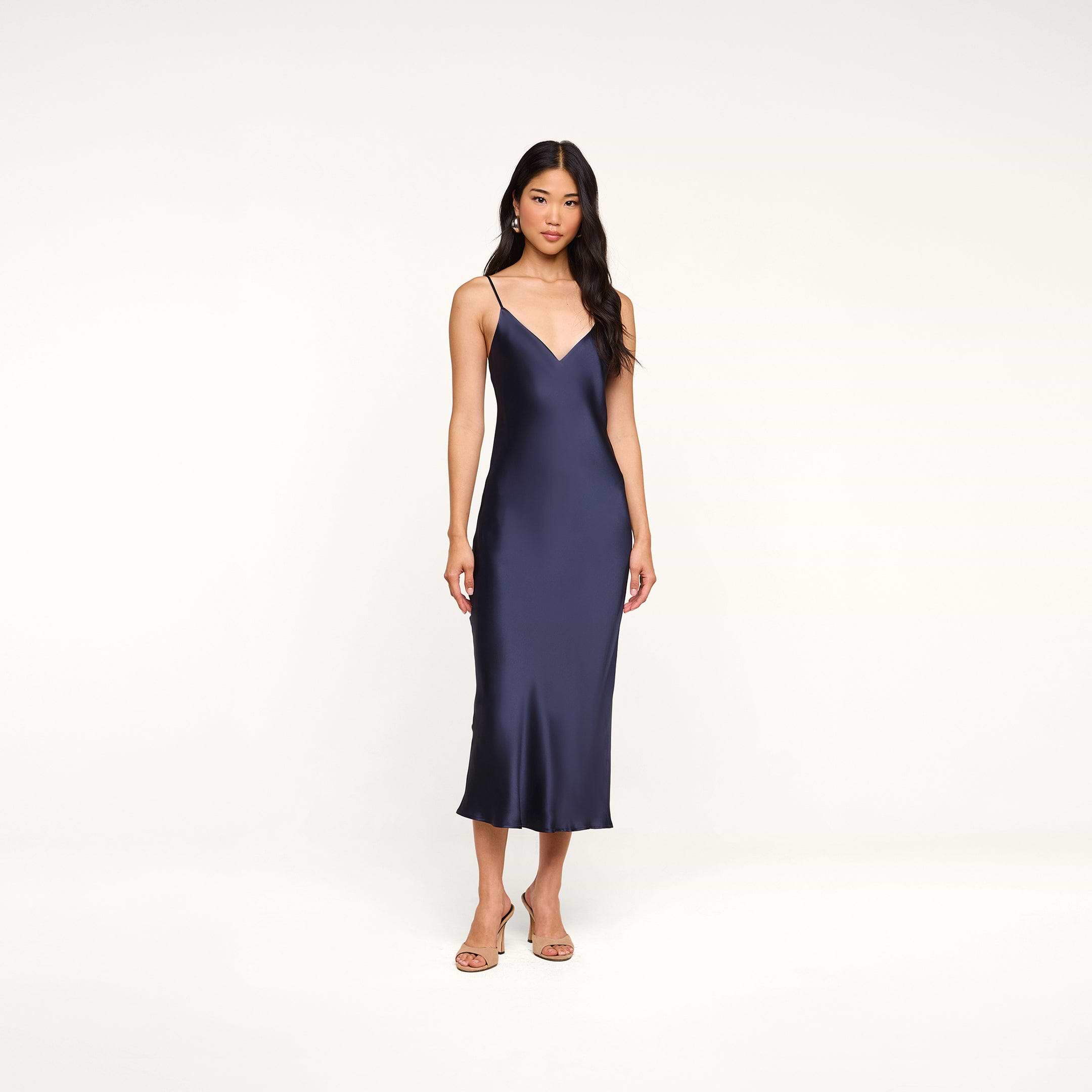 A woman stands against a plain white background wearing the Seridie Silk Slip Dress in navy blue with thin straps, paired with beige heeled sandals—an elegant choice for a wedding guest.