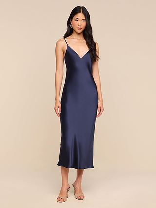 Wearing the Seridie Silk Slip Dress in navy blue with thin straps and paired with nude heels, this woman stands against a beige background—an ideal choice for an elegant wedding guest look.
